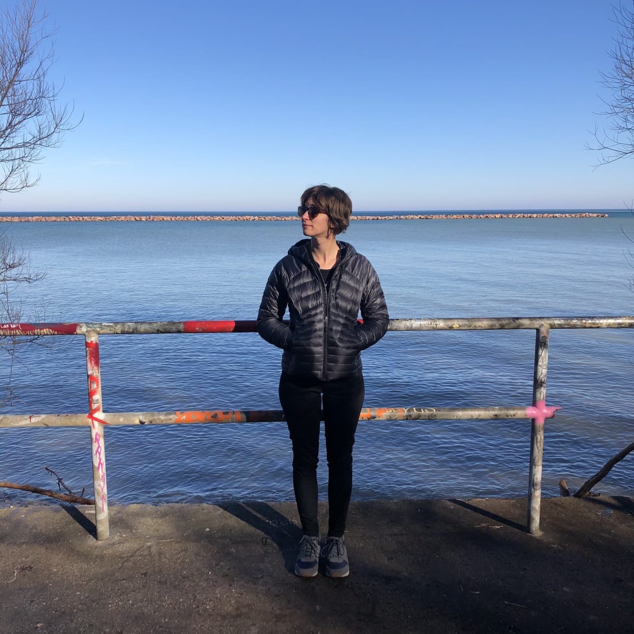 Mandi standing with her hands in her pocket up against a graffitied fence next. Lake Michigan and the break wall are in the background.