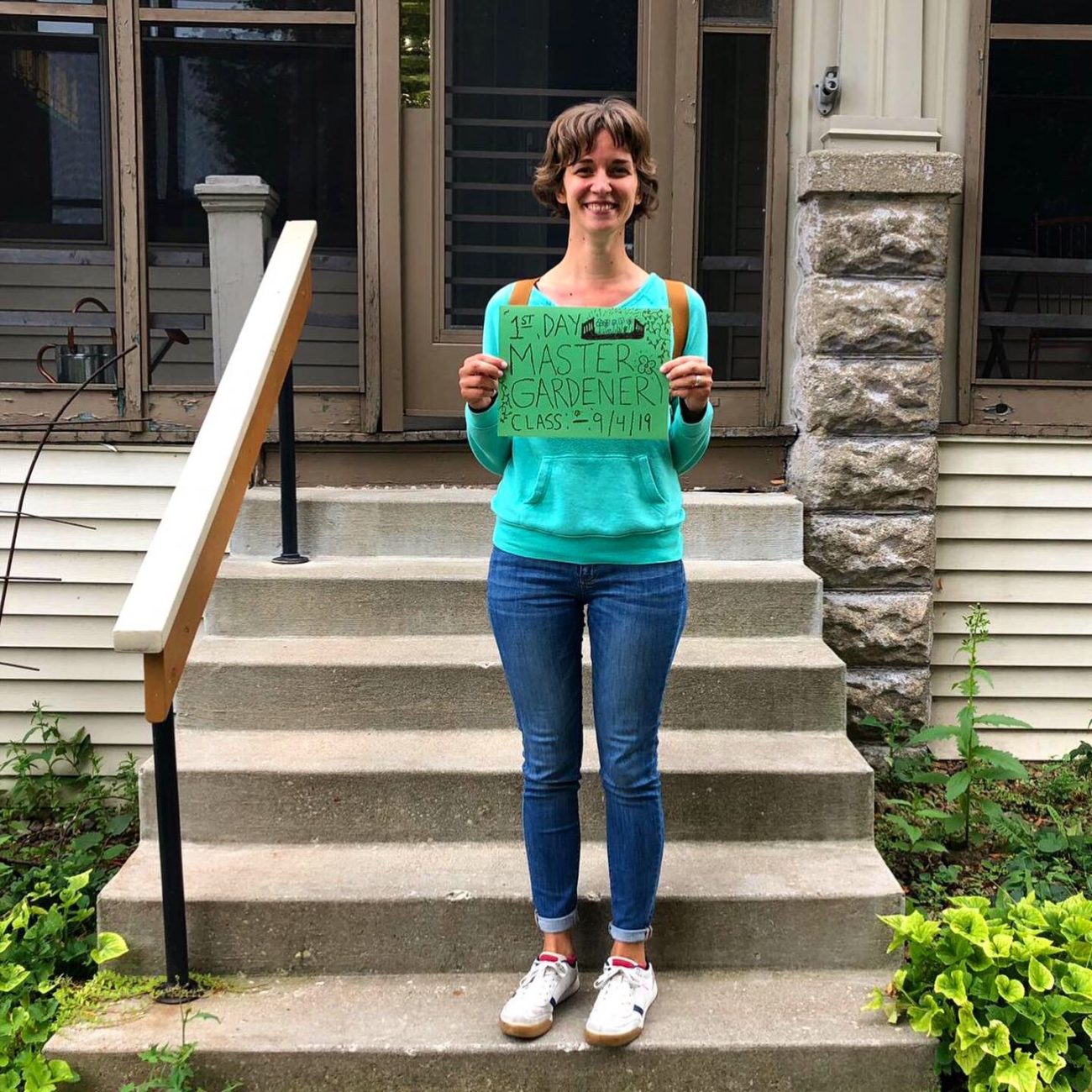 Mandi standing on her front porch with her backpack on. She's holding a sign that reads "1st Day Master Gardener Class - 9/4/19".