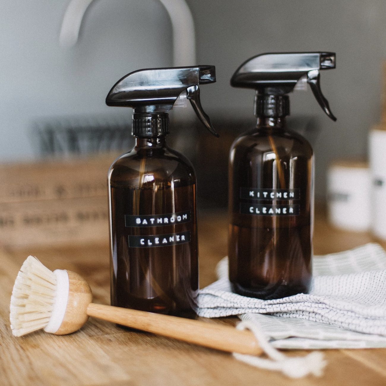 Two glass cleaning bottles sitting next to a wooden scrub brush and towels on a counter.