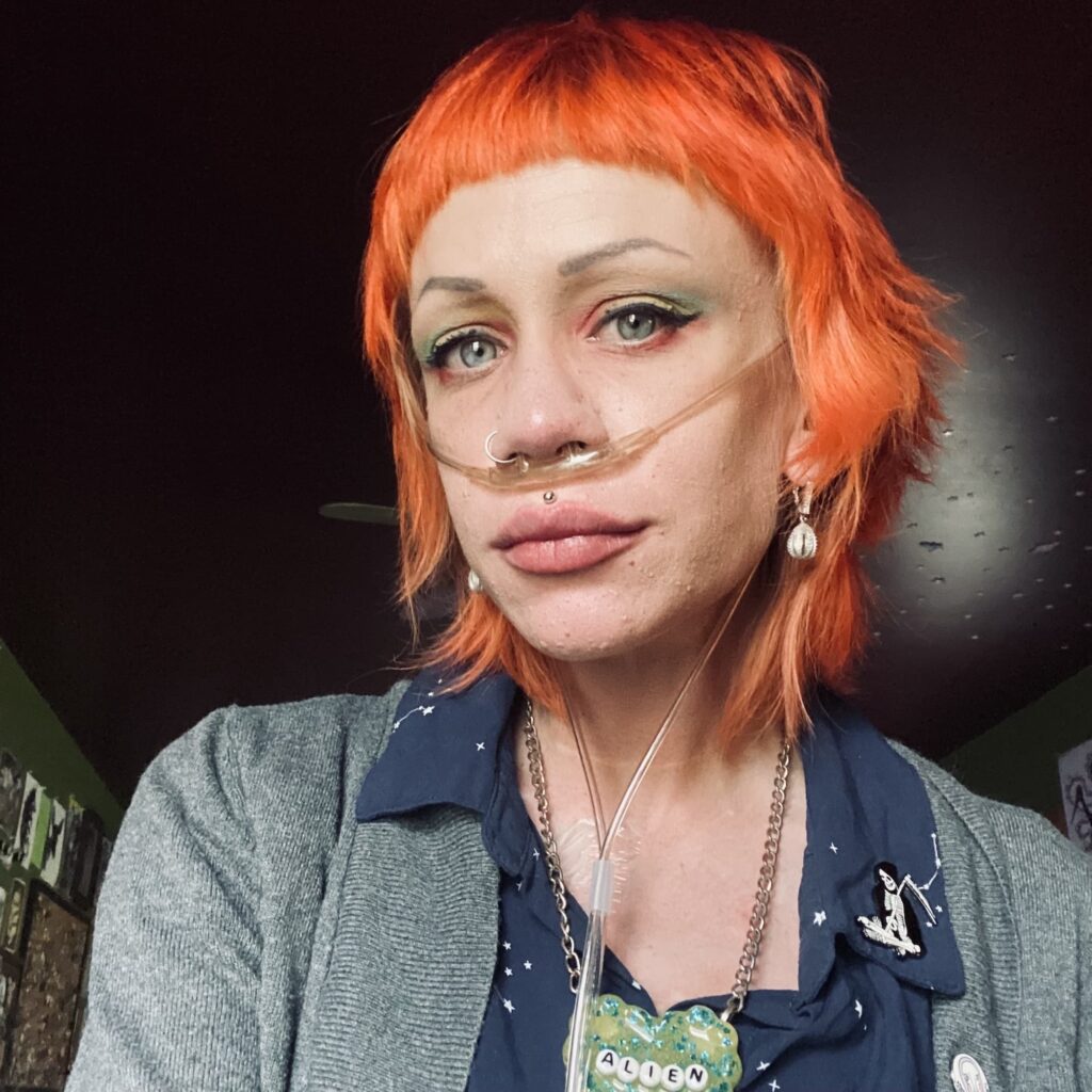 Jenny Janzer looking at the camera with a neutral gaze. She has chin length orange hair and is wearing oxygen tube, a necklace that says "alien" and a grey cardigan over a blue shirt.