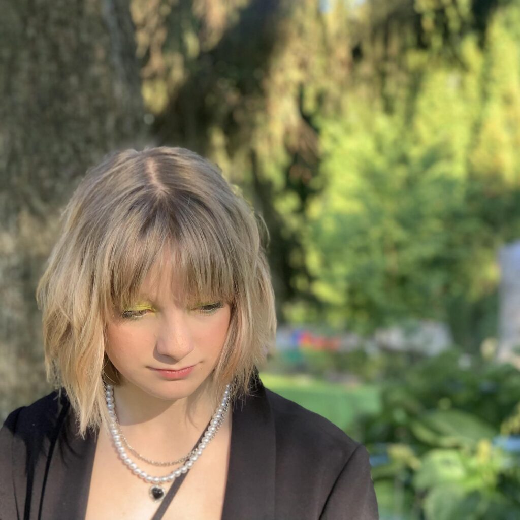 Lucia Sammarco with a neutral gaze looking towards the ground. They have blonde chin length hair and are wearing a necklace and black shirt.